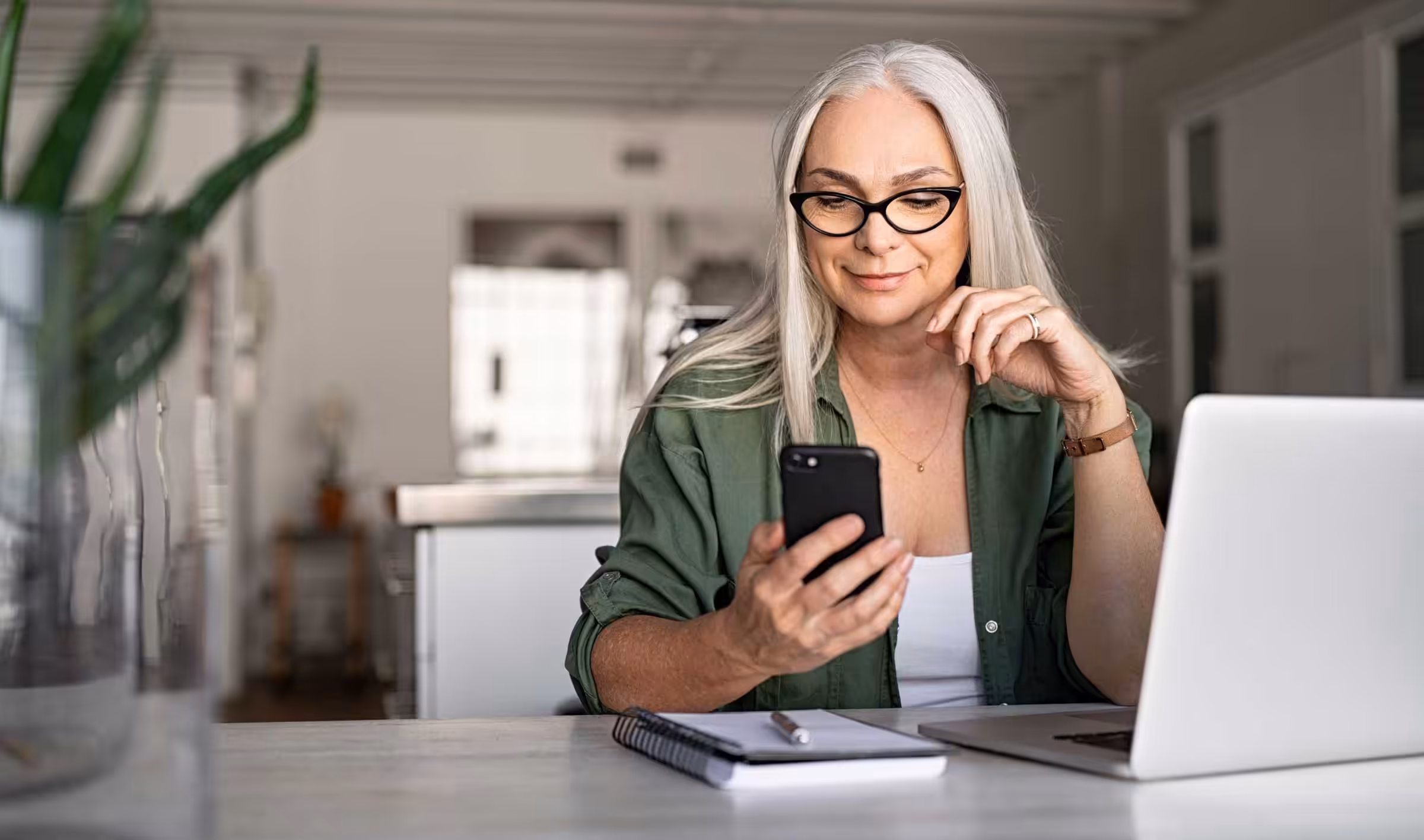 Happy-Woman-with-Smart-Phone-and-Laptop Happy senior woman using mobile phone while working at home with laptop. Smiling cool old woman wearing eyeglasses messaging with smartphone. Beautiful stylish elderly lady browsing site on cellphone.
