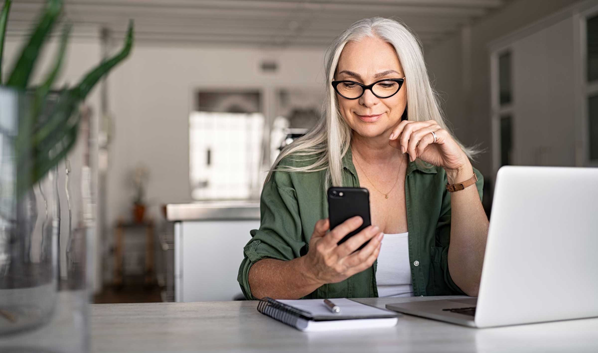 Happy senior woman using mobile phone while working at home with laptop. Smiling cool old woman wearing eyeglasses messaging with smartphone. Beautiful stylish elderly lady browsing site on cellphone.