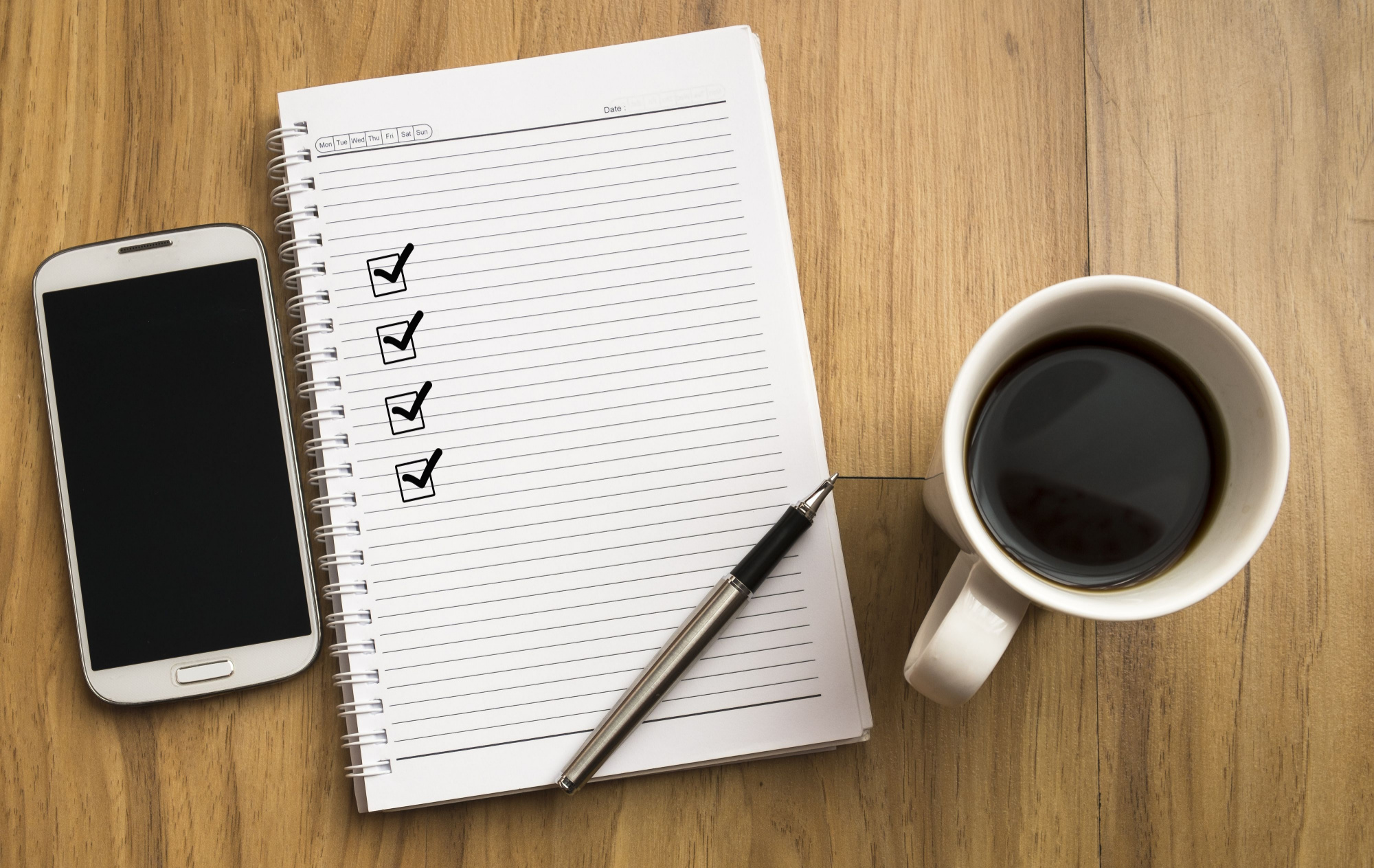 Note book with four check marks, smartphone, a pen and a cup of coffee on wooden background.