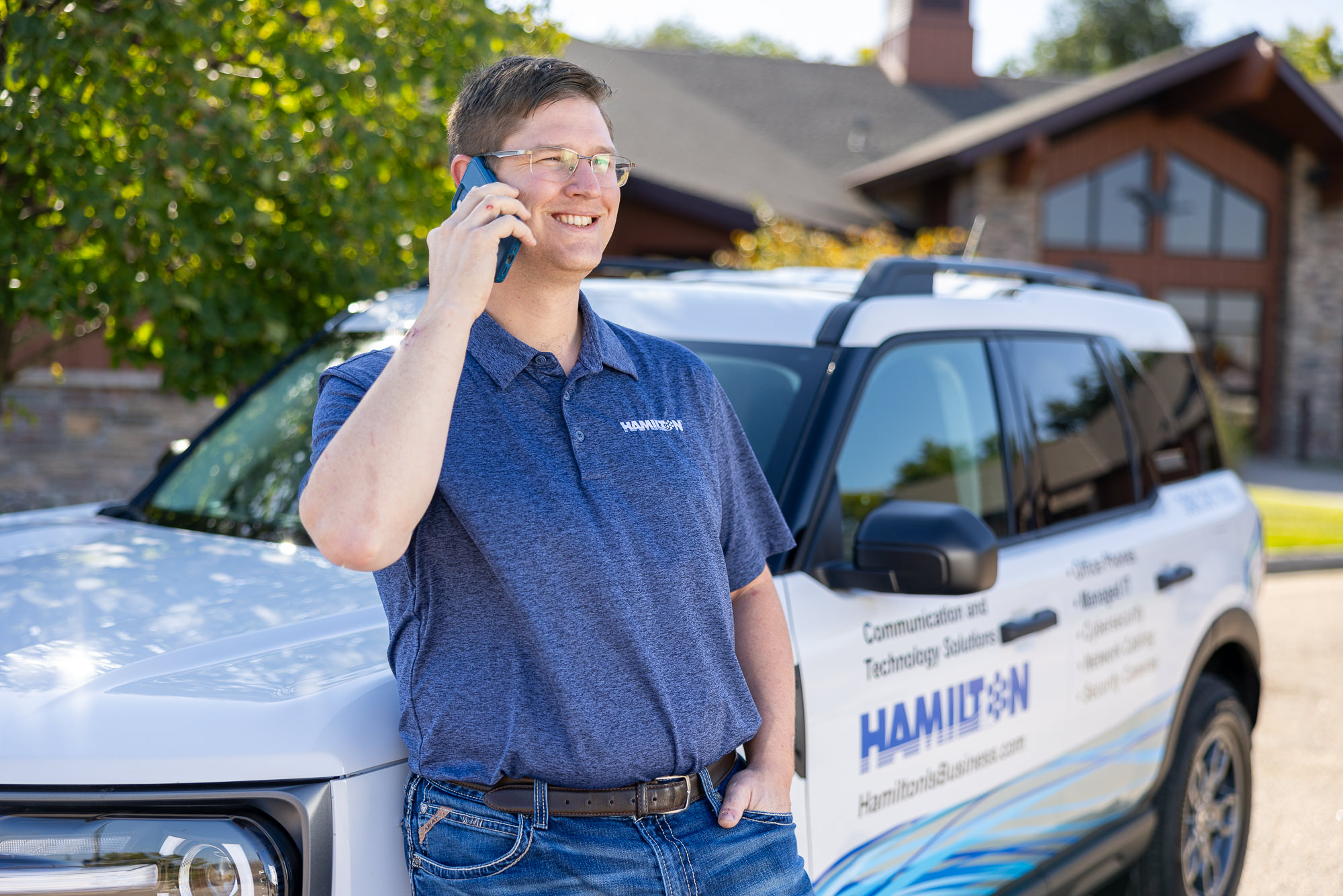 Hamilton employee talking on his modern mobile business phone system. leaning on his car with trees in the background